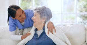 Caregiver smiling and comforting a senior woman wrapped in a blanket in a bright living room