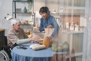 Caregiver unpacking groceries for a senior man in a wheelchair at a kitchen table