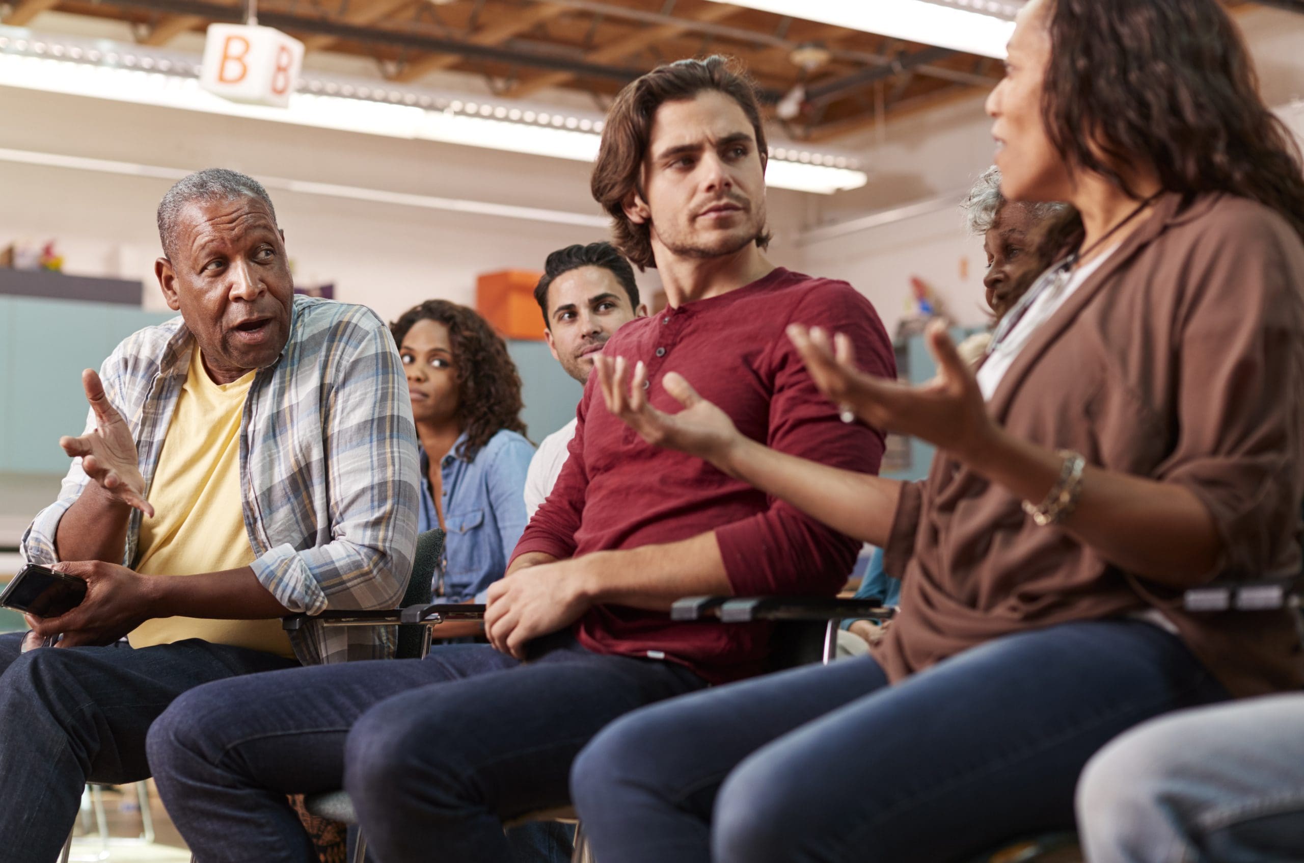 A mixed-age group of adults sits in a circle at a community center meeting in a bright room.